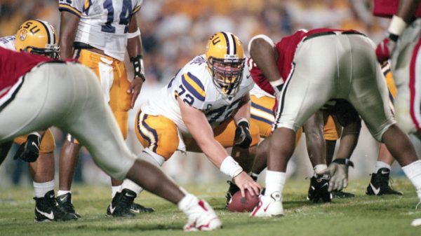 LSU offensive lineman snaps the ball at the line of scrimmage during a football play, surrounded by teammates in white and purple uniforms