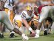 LSU offensive lineman snaps the ball at the line of scrimmage during a football play, surrounded by teammates in white and purple uniforms