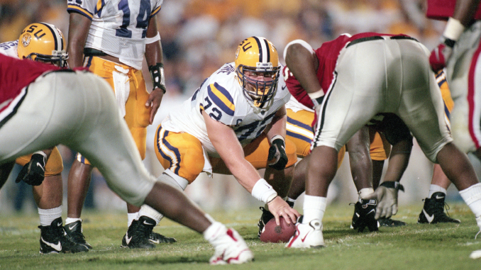 LSU offensive lineman snaps the ball at the line of scrimmage during a football play, surrounded by teammates in white and purple uniforms