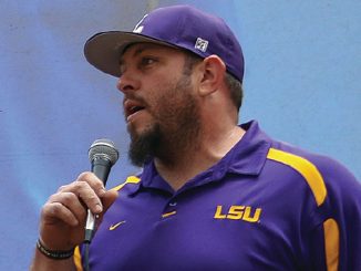 Man wearing a purple LSU polo shirt and cap, speaking into a handheld microphone at an event.