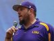 Man wearing a purple LSU polo shirt and cap, speaking into a handheld microphone at an event.