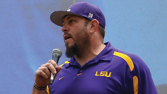 Man wearing a purple LSU polo shirt and cap, speaking into a handheld microphone at an event.