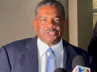 Man in a dark pinstripe suit and sky-blue tie speaks to reporters, microphones in front of him outside a building.