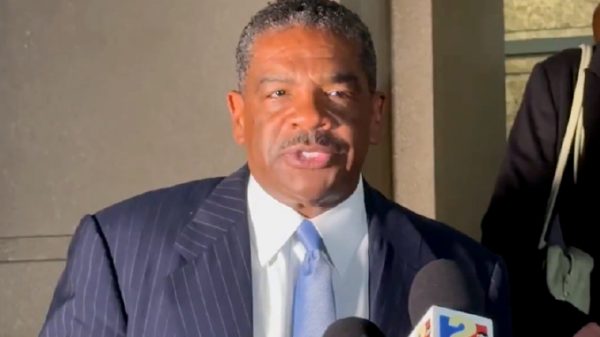 Man in a dark pinstripe suit and sky-blue tie speaks to reporters, microphones in front of him outside a building.