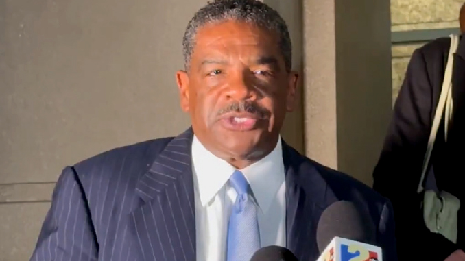 Man in a dark pinstripe suit and sky-blue tie speaks to reporters, microphones in front of him outside a building.