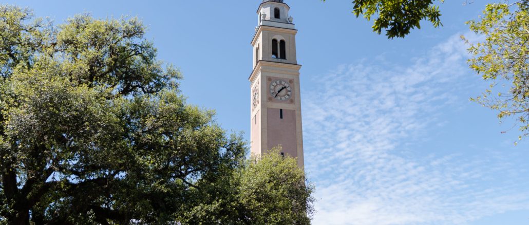 Clock tower rising above a beige building, framed by green trees under a clear blue sky.