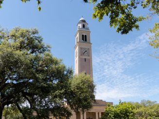 Clock tower rising above a beige building, framed by green trees under a clear blue sky.