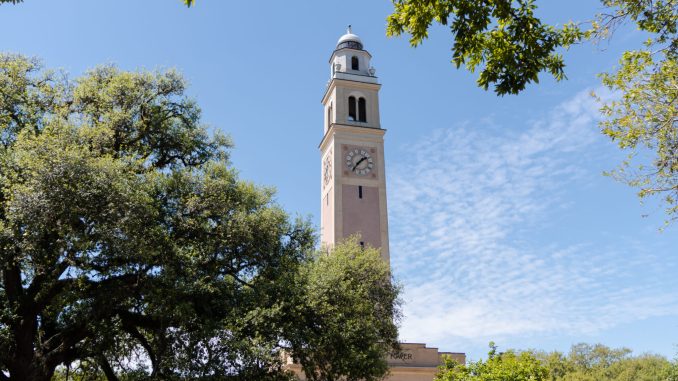 Clock tower rising above a beige building, framed by green trees under a clear blue sky.
