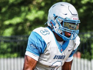 Football player in BYNES white and light blue uniform crouches on the field, wearing a blue helmet with a facemask.