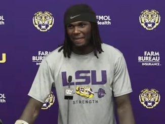LSU football player stands at a podium during a press conference, wearing a gray LSU shirt with 'STRENGTH' on it against a purple backdrop with logos.