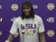 LSU football player stands at a podium during a press conference, wearing a gray LSU shirt with 'STRENGTH' on it against a purple backdrop with logos.