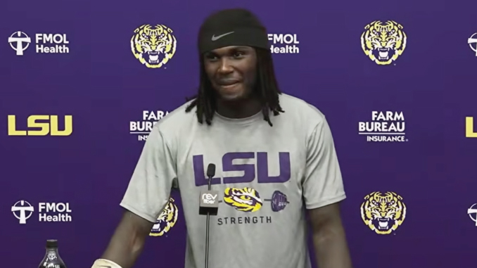 LSU football player stands at a podium during a press conference, wearing a gray LSU shirt with 'STRENGTH' on it against a purple backdrop with logos.