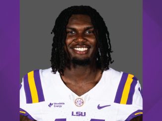 Portrait of an LSU football player smiling, wearing a white jersey with purple and gold stripes and LSU logo.