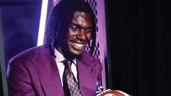 Smiling man in a purple suit and patterned tie, holding a basketball at an indoor event with purple lighting behind him