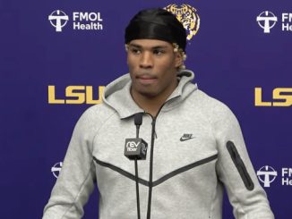 Male athlete in a gray Nike hoodie and black headscarf speaking at a LSU press conference, microphone in front, purple backdrop with LSU logos.