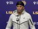 Male athlete in a gray Nike hoodie and black headscarf speaking at a LSU press conference, microphone in front, purple backdrop with LSU logos.