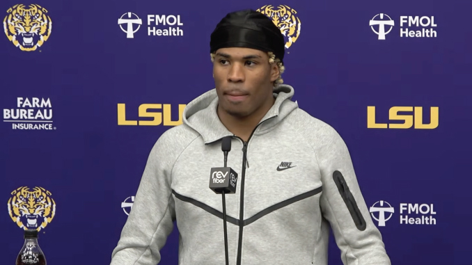 Male athlete in a gray Nike hoodie and black headscarf speaking at a LSU press conference, microphone in front, purple backdrop with LSU logos.