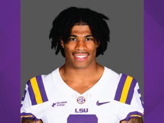 Portrait of a college football player in an LSU jersey, smiling at the camera against a gray backdrop.