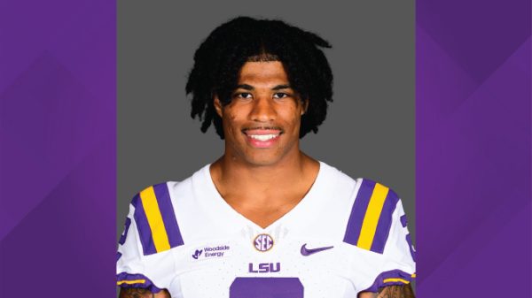 Portrait of a college football player in an LSU jersey, smiling at the camera against a gray backdrop.