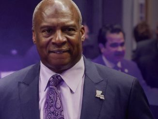 Smiling man in a dark suit and purple paisley tie at a formal event, wearing a US-shaped pin.