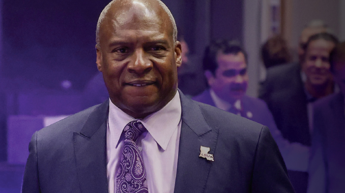 Smiling man in a dark suit and purple paisley tie at a formal event, wearing a US-shaped pin.