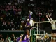 Female volleyball player in purple leaps to spike the ball over the net as the crowd watches from stands behind her.
