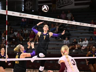 LSU volleyball player leaps to spike the ball over the net as teammates watch nearby.