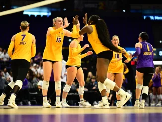 Female volleyball players in yellow uniforms high-fiving on the court during a match, with a purple-clad opponent nearby and spectators in the background.