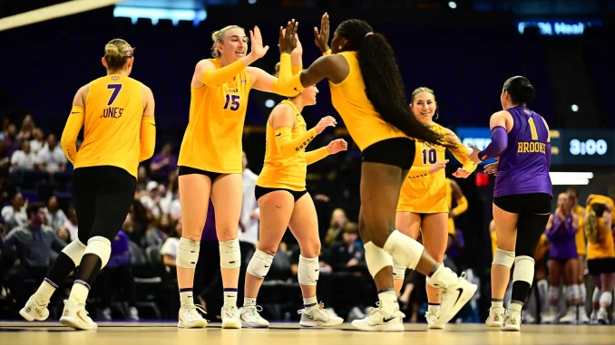 Female volleyball players in yellow uniforms high-fiving on the court during a match, with a purple-clad opponent nearby and spectators in the background.