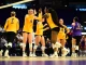Female volleyball players in yellow uniforms high-fiving on the court during a match, with a purple-clad opponent nearby and spectators in the background.