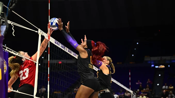 Female volleyball players leap at the net, reaching for a blue-and-white ball during a match.