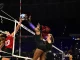 Female volleyball players leap at the net, reaching for a blue-and-white ball during a match.
