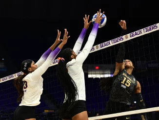 Volleyball players jump near the net for a block; two in white reach up as a ball sails above them, while a dark-uniform opponent attempts a spike.