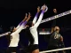 Volleyball players jump near the net for a block; two in white reach up as a ball sails above them, while a dark-uniform opponent attempts a spike.