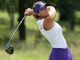 Female golfer in a white sleeveless top and purple visor swinging a driver on a green golf course as trees blur in the background.