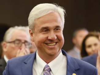 Smiling man in a blue suit and lavender tie at a social event with people in the background.