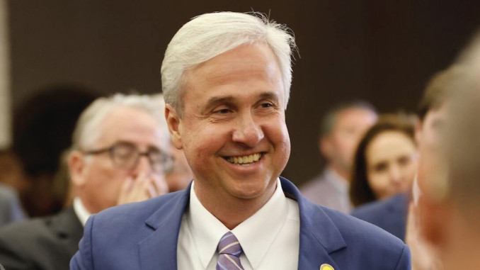 Smiling man in a blue suit and lavender tie at a social event with people in the background.