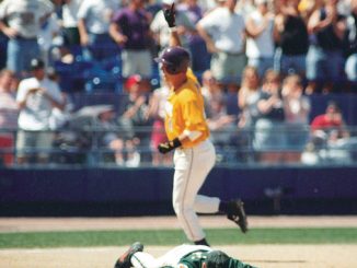 Baseball player in a yellow uniform runs past a fallen catcher on the infield as fans watch from the stands.