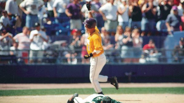 Baseball player in a yellow uniform runs past a fallen catcher on the infield as fans watch from the stands.