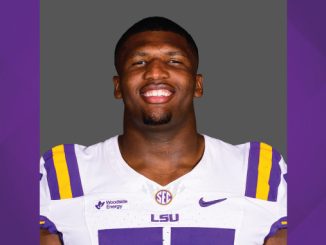 Portrait of an LSU football player smiling, wearing a white jersey with purple and gold stripes and LSU branding.