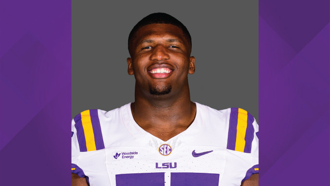 Portrait of an LSU football player smiling, wearing a white jersey with purple and gold stripes and LSU branding.