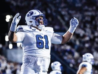 American football player wearing #61 in a white BYU uniform, raising his arms in celebration on the field with a cheering crowd behind him.