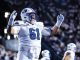 American football player wearing #61 in a white BYU uniform, raising his arms in celebration on the field with a cheering crowd behind him.