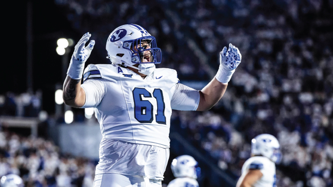 American football player wearing #61 in a white BYU uniform, raising his arms in celebration on the field with a cheering crowd behind him.