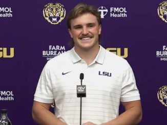 Portrait of a smiling man in a white LSU polo at a podium during a press conference, with a purple backdrop featuring LSU logos and branding.