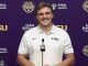 Portrait of a smiling man in a white LSU polo at a podium during a press conference, with a purple backdrop featuring LSU logos and branding.