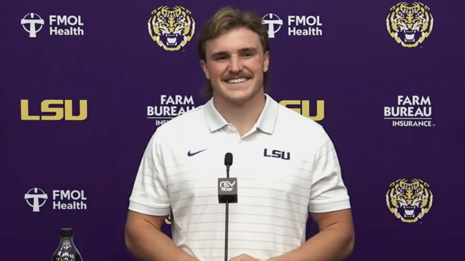 Portrait of a smiling man in a white LSU polo at a podium during a press conference, with a purple backdrop featuring LSU logos and branding.