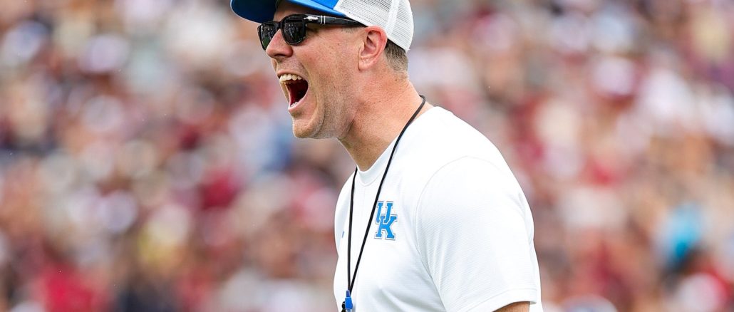 Coach wearing a white shirt, blue cap, sunglasses, and a whistle on a lanyard during a game.