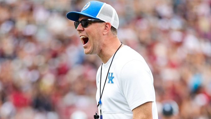 Coach wearing a white shirt, blue cap, sunglasses, and a whistle on a lanyard during a game.