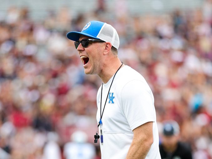 Coach wearing a white shirt, blue cap, sunglasses, and a whistle on a lanyard during a game.